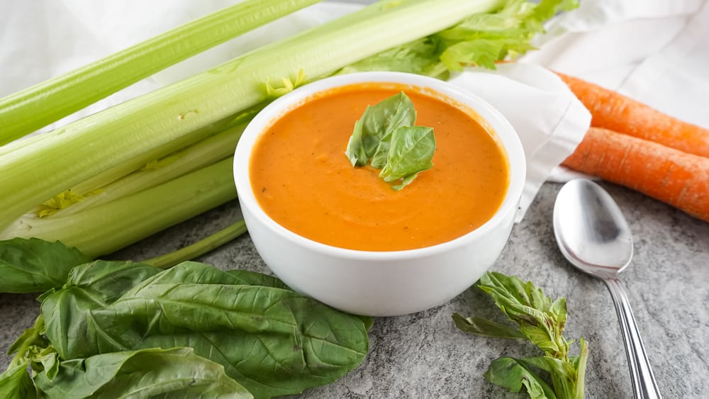 Homemade copycat Panera tomato soup garnished with basil, with celery and carrots in the background