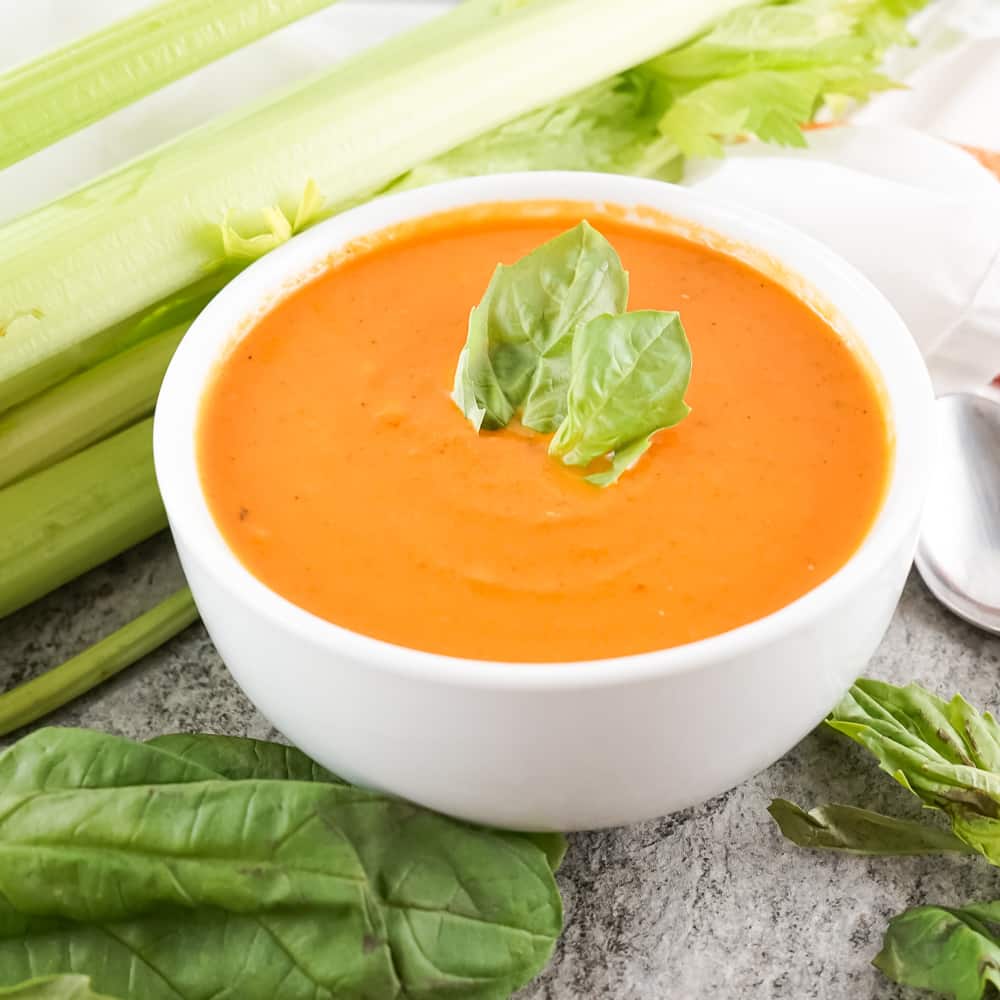 Creamy tomato soup in a white bowl with basil leaves, celery and carrots