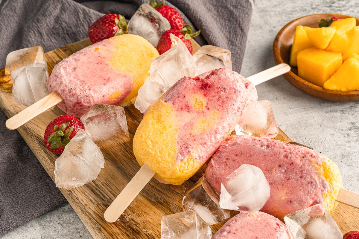 strawberry mango popsicles stacked on wooden platter with ice cubes