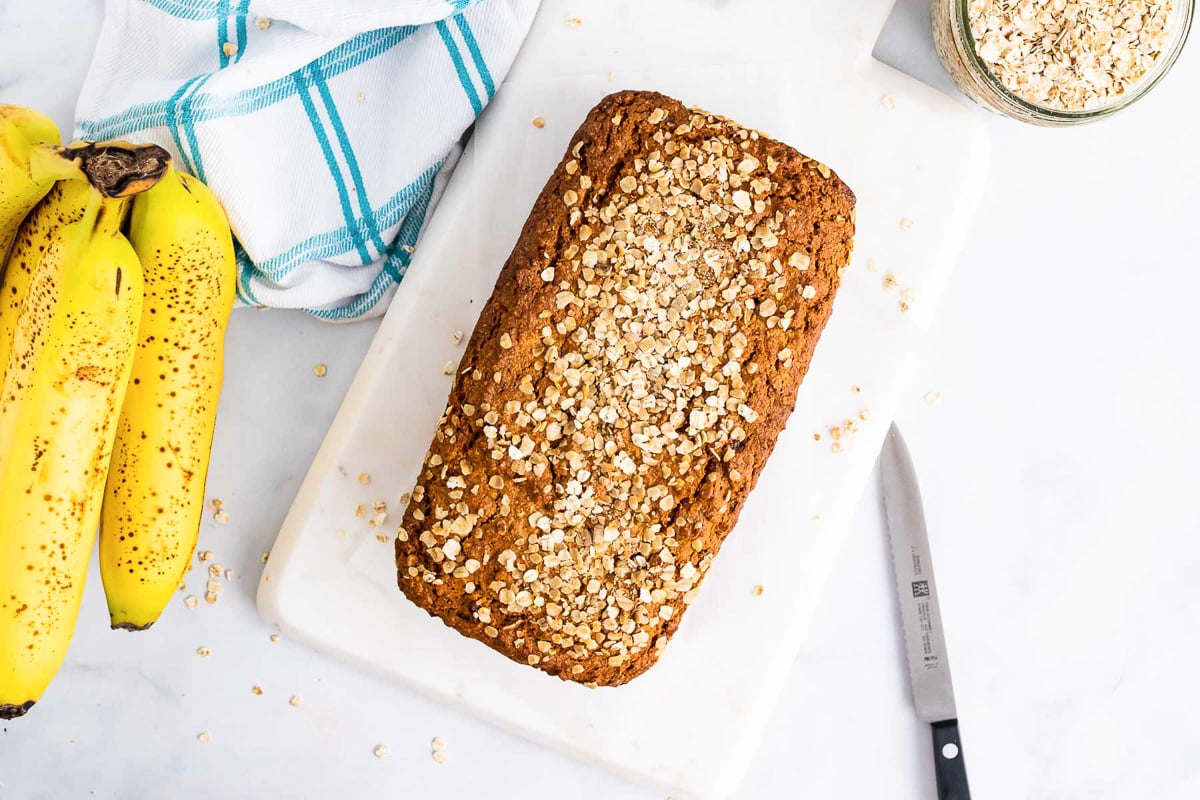 loaf of banana oat bread on white counter next to slicing knife and bunch of bananas