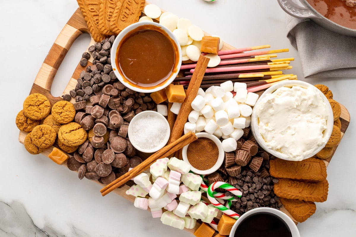 top view of hot chocolate charcuterie board with cookies, sauce, whipped cream, pokeys, marshmallows, candy canes, chocolate chips, and caramels