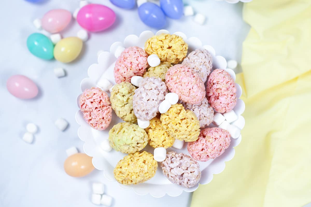 top view of easter rice krispie treats on white serving platter sprinkled with white mini marshmallows and plastic Easter eggs in the background