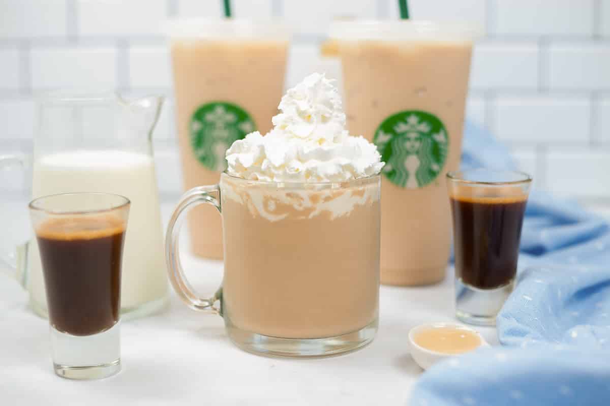 side view of copycat starbucks white chocolate mocha in clear mug surrounded by ingredients to make it and Starbucks tumblers