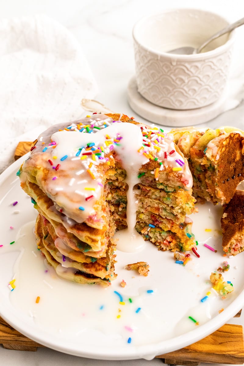 stack of funfetti pancakes with slice cut out topped with icing and served on white plate