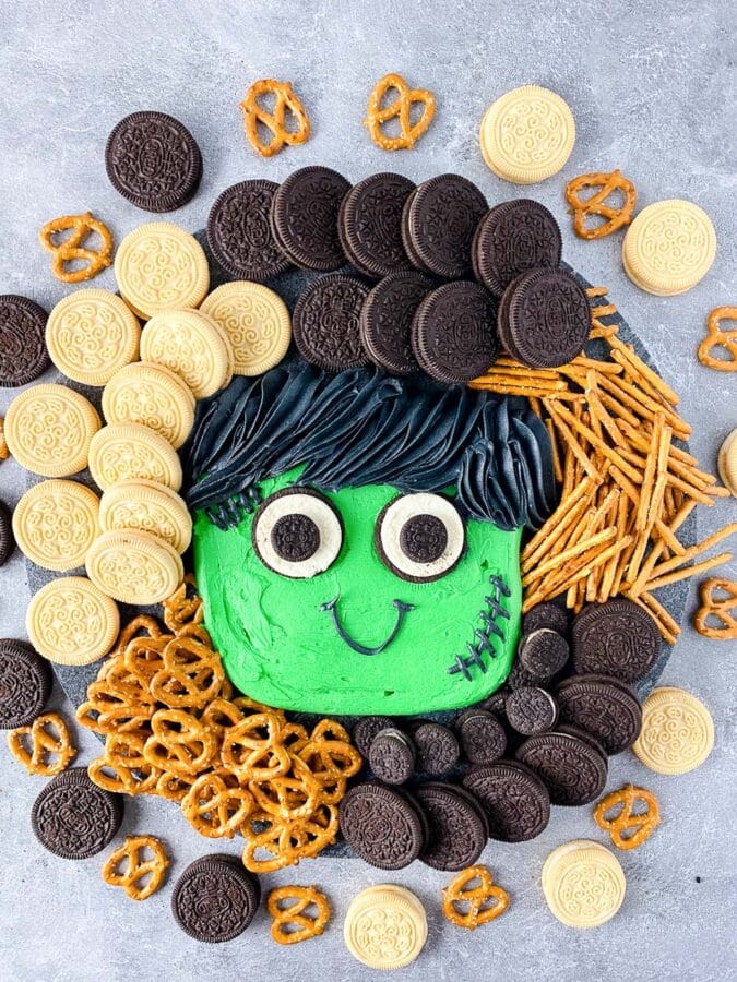 Halloween buttercream board with a green Frankenstein face, sandwich cookies and pretzels on a slate board