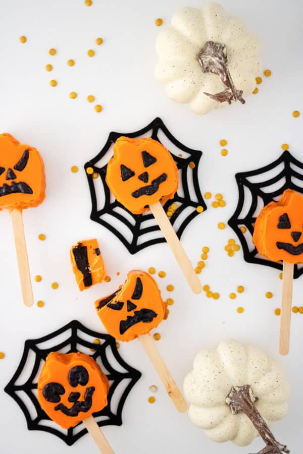 Orange jack-o'-lantern cake pops on sticks arranged on black spiderweb coasters with white mini pumpkins