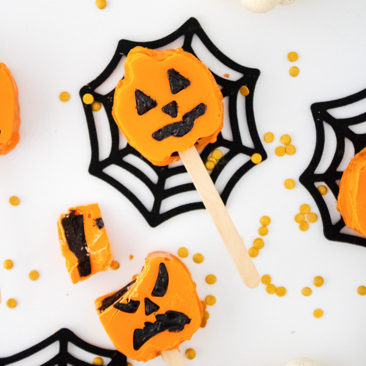 Close-up of a jack-o'-lantern cake pop on a black spiderweb coaster next to a cut pop showing the chocolate cake inside