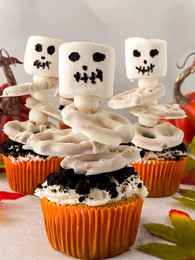 Halloween skeleton cupcakes with marshmallow skulls and white candy-coated pretzel ribcages, surrounded by mini pumpkins and fall leaves