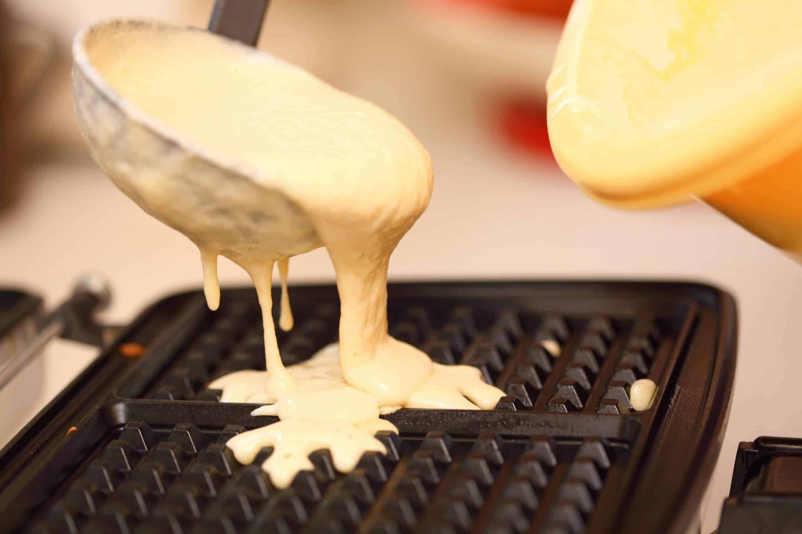 Waffle batter overflowing from a ladle onto a hot waffle iron griddle