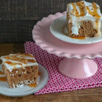 Two slices of apple spice poke cake topped with whipped topping, caramel and chopped nuts, one on a pink cake stand.