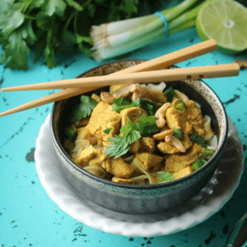 Chicken curry with noodles in a dark bowl topped with cashews and herbs, with chopsticks