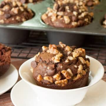 Chocolate chocolate chip banana nut muffin topped with chopped walnuts in a white cup, with more muffins in a jumbo muffin pan behind