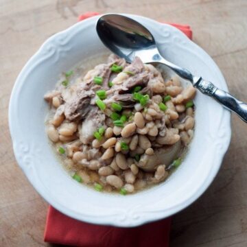 Crock pot ham and white beans in a white bowl with a spoon, topped with chopped chives