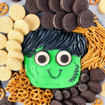 Halloween buttercream board with a green Frankenstein face surrounded by chocolate and vanilla sandwich cookies and pretzels