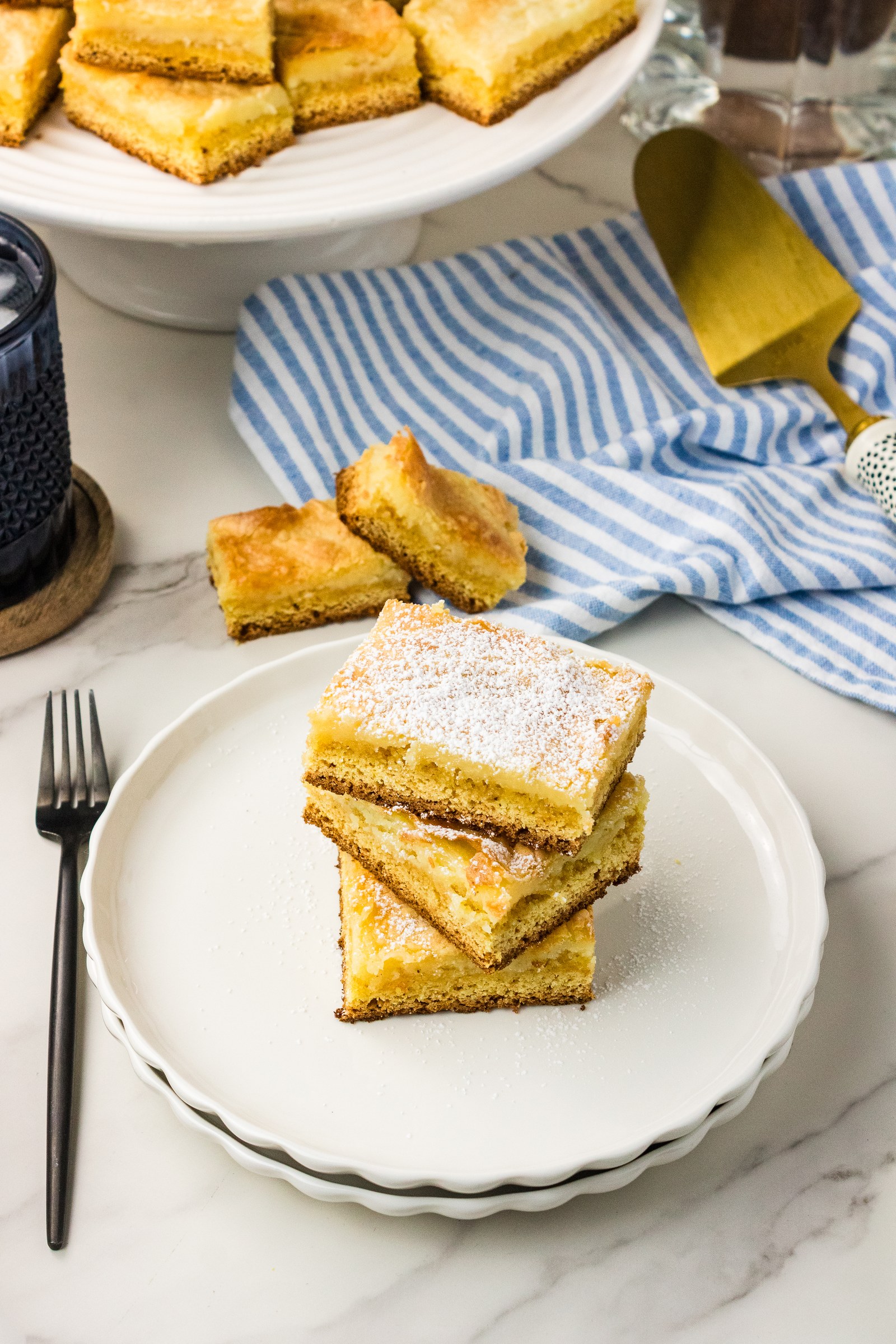Stack of gooey butter cake bars on a white plate with a fork and more bars on a cake stand behind