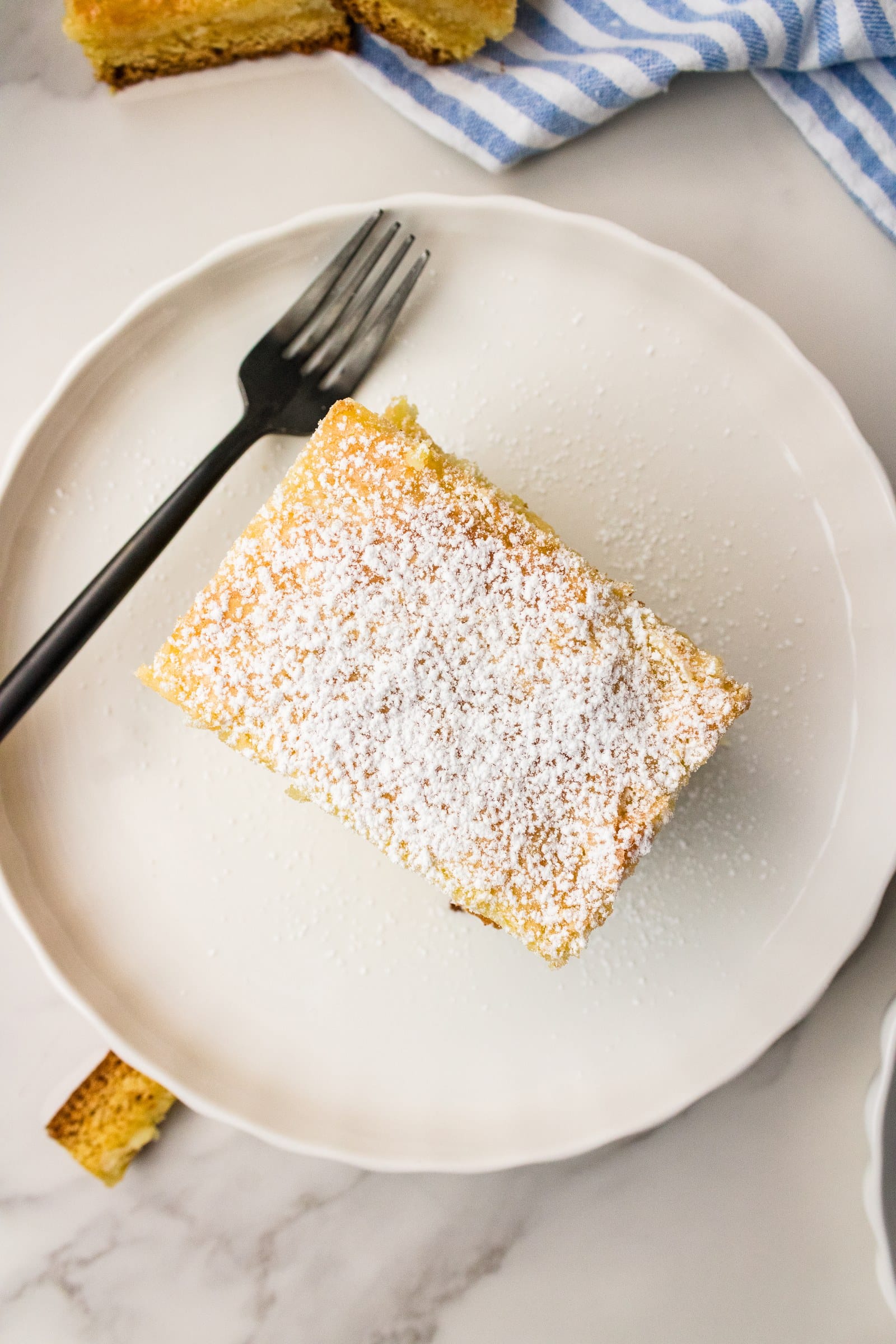 Overhead view of a square of gooey butter cake dusted with powdered sugar on a white plate with a fork