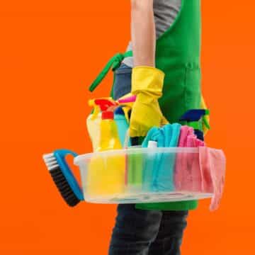 Person in a green apron and yellow rubber gloves carrying a caddy of cleaning supplies against an orange background
