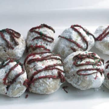 Peppermint snowball cookies dusted in powdered sugar with a chocolate drizzle and red and green sprinkles