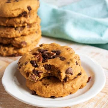 Two pumpkin chocolate chip cookies on a white plate with a stack of cookies behind them