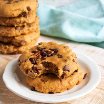 Two soft pumpkin chocolate chip cookies with pecans on a small white plate, one with a bite taken out