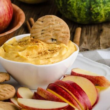 Pumpkin cream cheese filling in a white bowl dusted with cinnamon, with a cookie and fresh apple slices