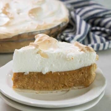 Slice of pumpkin meringue pie with a tall toasted meringue topping on a white plate, with the whole pie and a striped towel behind it