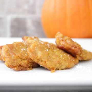 Golden fried pumpkin patties dusted with powdered sugar on a white plate with a whole pumpkin behind them