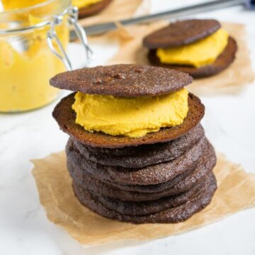 Stack of pumpkin whoopie pies with pumpkin cream filling on parchment paper, with a jar of pumpkin cream behind