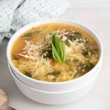 Square photo of roasted tomato and cauliflower rice soup with kale, shredded Parmesan and a basil leaf in a white bowl
