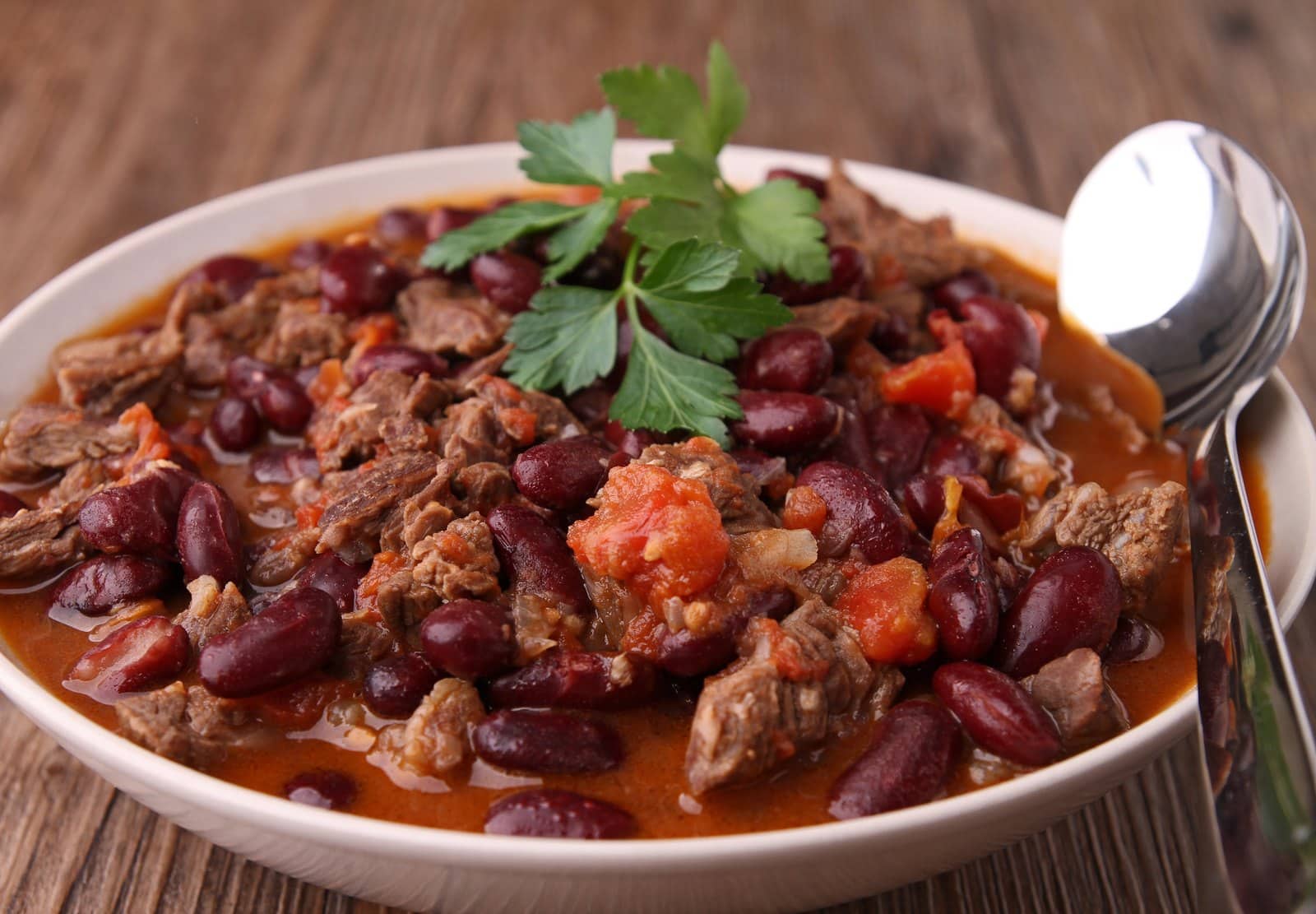 Bowl of healthy beef and kidney bean chili garnished with parsley, with a spoon on a wooden table