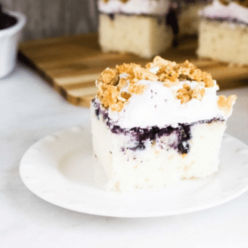 A slice of blueberry poke cake with whipped topping and graham crumble on a white plate
