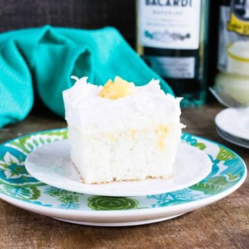 Slice of boozy pina colada poke cake topped with whipped topping, coconut and pineapple on a patterned plate