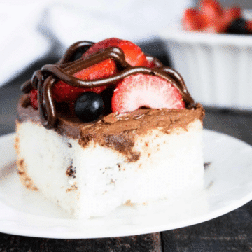 Slice of chocolate ganache poke cake topped with whipped ganache, strawberries, blueberries and a chocolate drizzle on a white plate