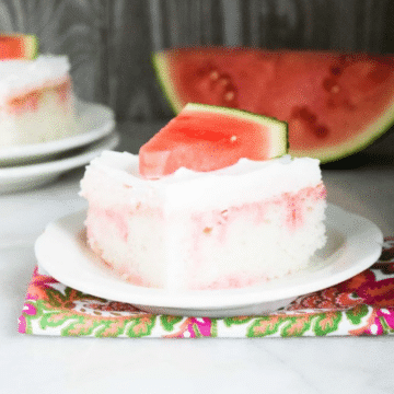 Slice of Jolly Rancher watermelon poke cake with white frosting and a fresh watermelon wedge on a white plate