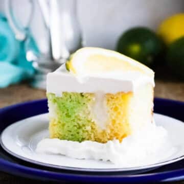 Slice of lemon lime poke cake with green and yellow gelatin stripes, whipped topping and a lemon slice on a blue plate