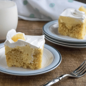 Two slices of pineapple poke cake with vanilla pudding and whipped topping on blue-rimmed plates, with a glass of milk